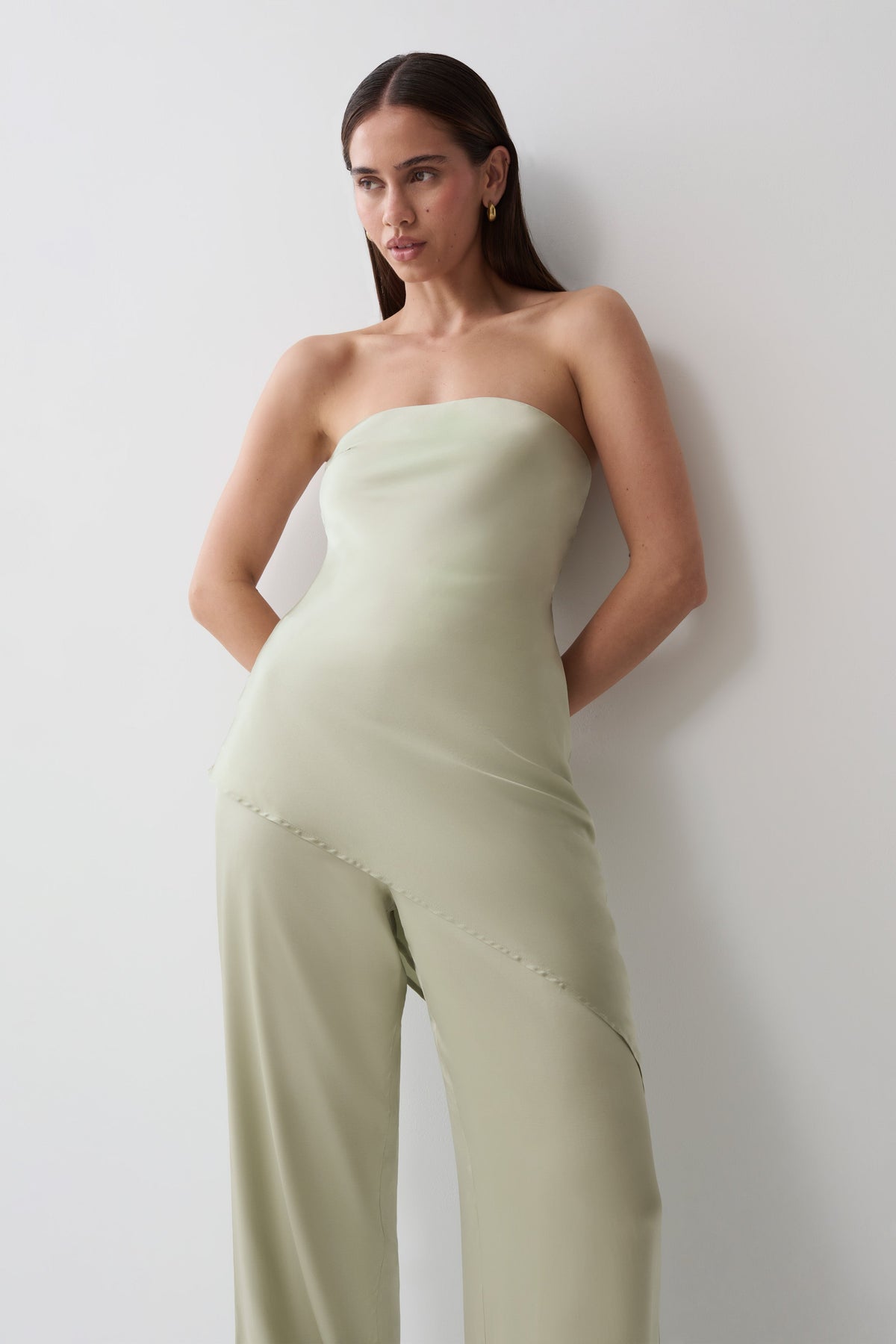 A woman wearing a strapless sage green strapless top with wide-leg pants, posing against a plain light gray wall.
