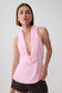 A woman modeling a light pink sleeveless top with a deep ruffled cowl neckline, paired with dark brown shorts, against a plain light gray studio background.