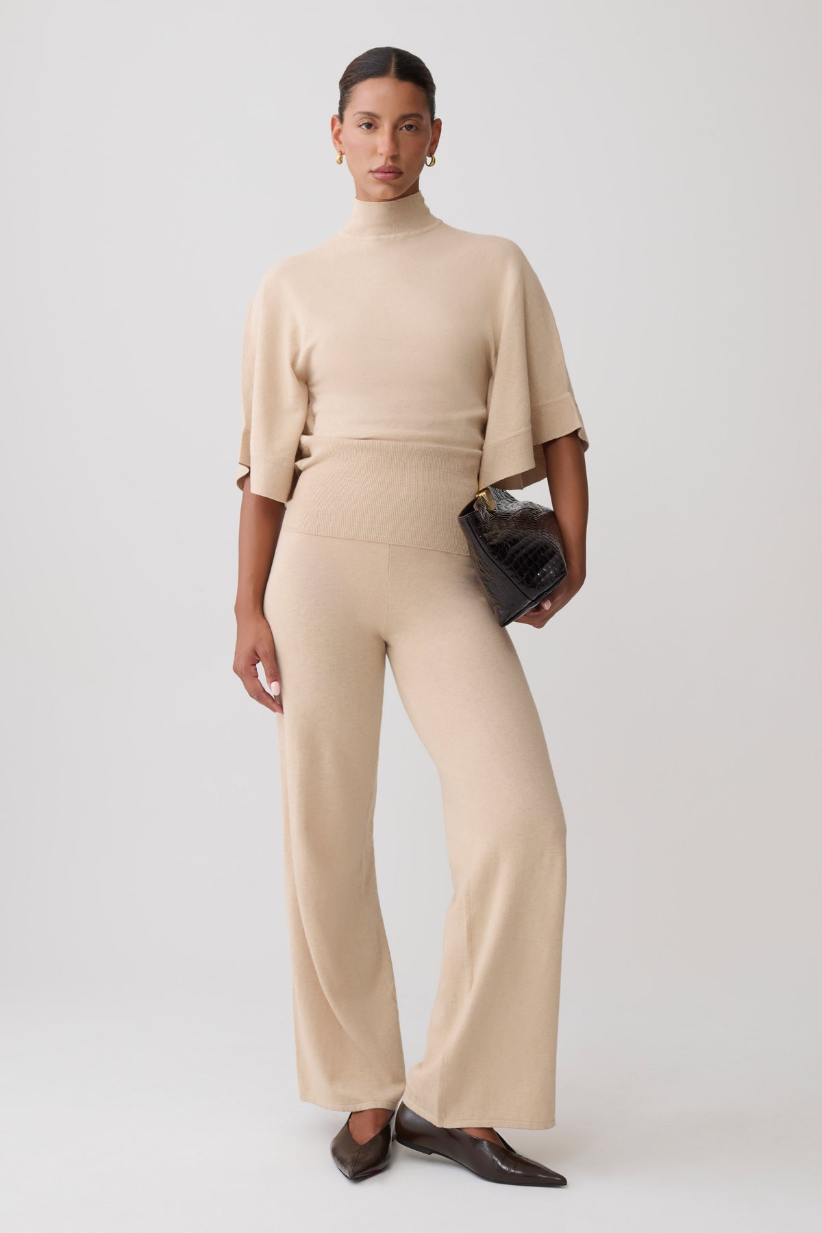 A model stands against a light gray studio background wearing a matching beige knit two-piece set consisting of a short-sleeve mock-neck top and wide-leg trousers, paired with dark brown pointed flats and a small black textured clutch.