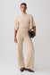 A model stands against a light gray studio background wearing a matching beige knit two-piece set consisting of a short-sleeve mock-neck top and wide-leg trousers, paired with dark brown pointed flats and a small black textured clutch.