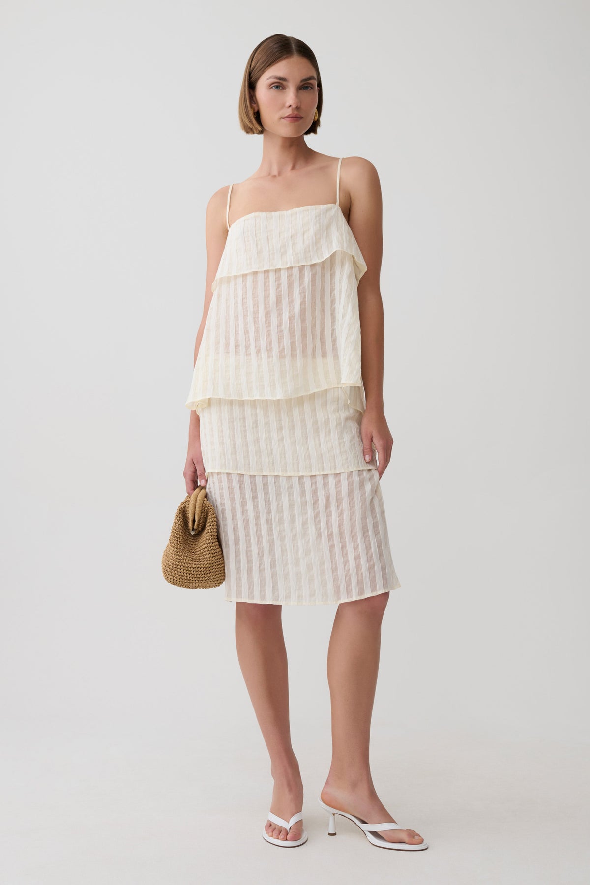 A woman models a cream-colored tiered top with thin spaghetti straps and subtle vertical stripes and matching midi skirt, styled with a small woven straw bucket bag and white slide sandals against a light gray studio background.