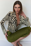 A model wearing a black and beige zebra-print long-sleeve top and textured olive green pants, posing against a plain white wall.