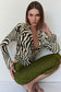 A model wearing a black and beige zebra-print long-sleeve top and textured olive green pants, posing against a plain white wall.