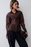 A woman models a sheer, long-sleeved button-up blouse in a dark brown color, paired with dark wide-leg trousers and holding a black textured clutch bag against a plain light gray background.