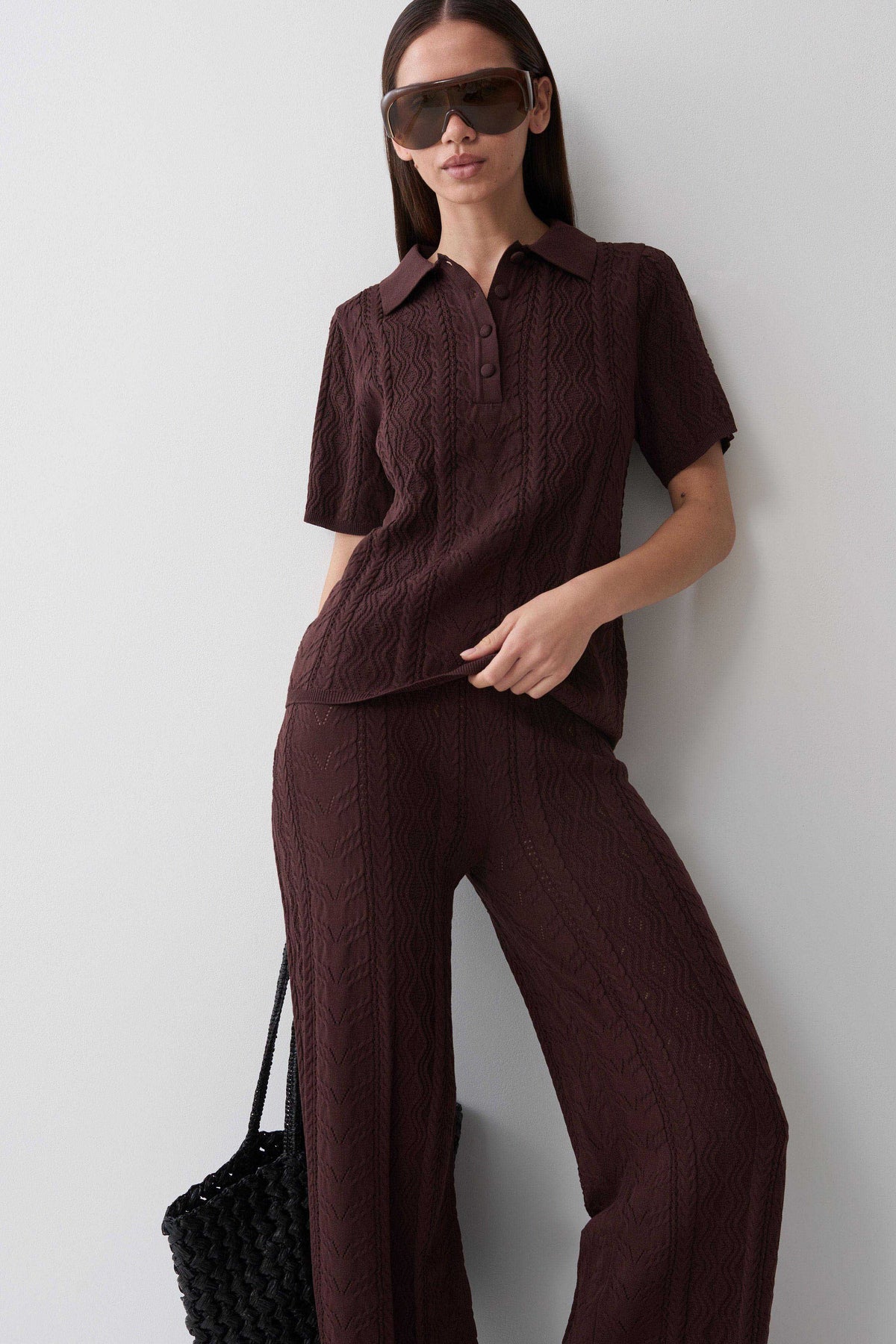 A woman models a matching dark brown cable-knit two-piece set consisting of a short-sleeve collared polo shirt and wide-leg trousers, styled with oversized sunglasses and a black woven handbag against a plain light gray wall.