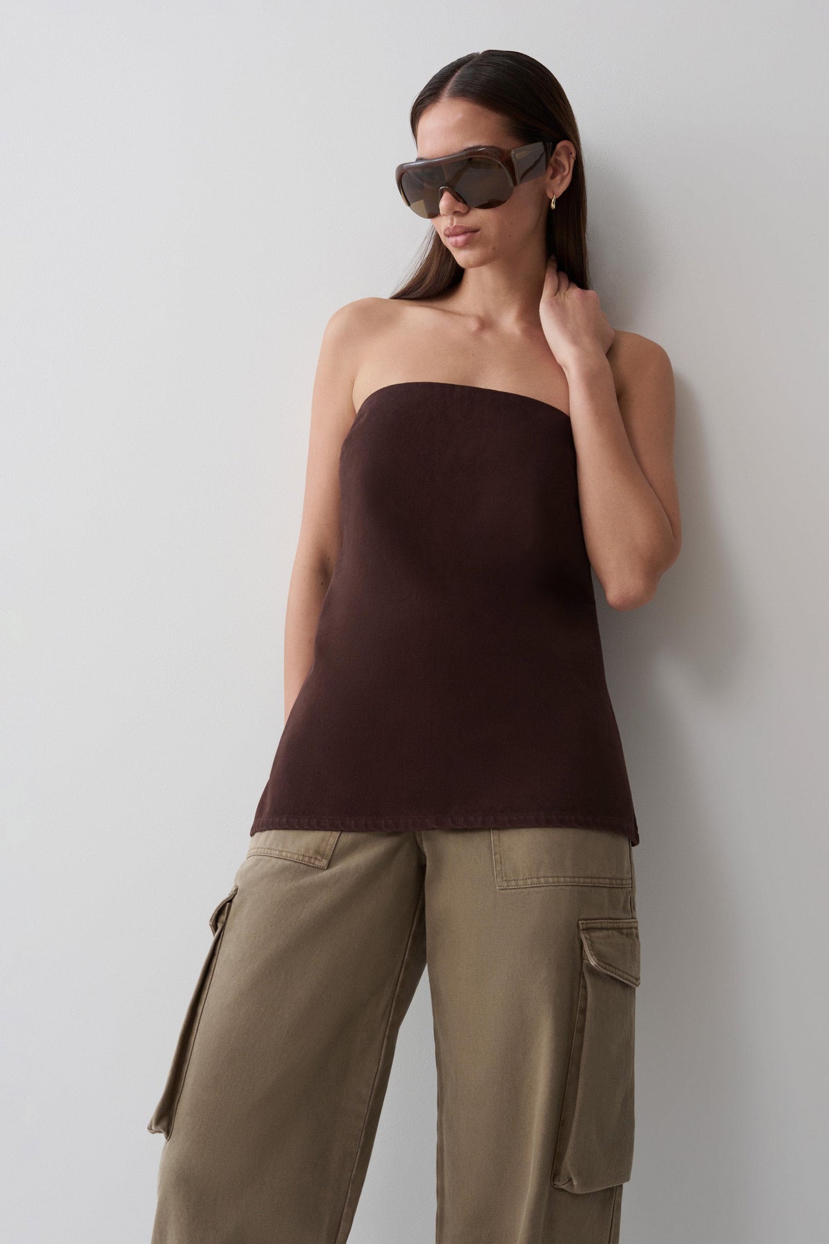 A woman wearing a dark brown strapless tube top and khaki cargo pants, standing against a light gray wall.