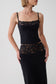 A woman wearing a black camisole-style top with floral lace trim at the neckline and waist, paired with a long black skirt, against a light gray studio background.
