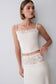 A woman modeling a cream-colored two-piece set consisting of a spaghetti-strap crop top with lace trim and a matching high-waisted skirt with lace detailing, set against a plain light gray background.