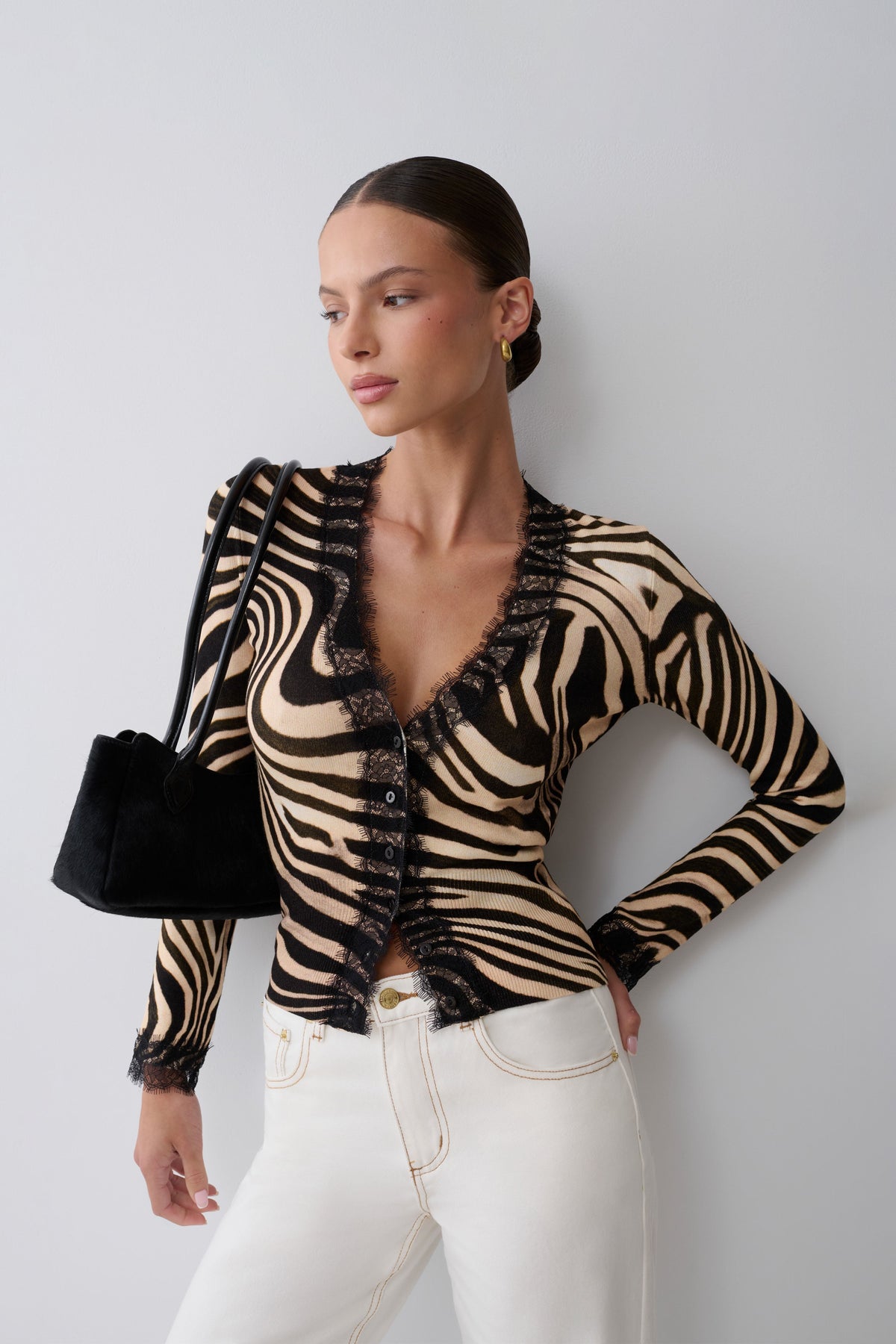 A woman modeling a beige and black zebra-print long-sleeve top with black lace trim, paired with white jeans and holding a black handbag against a light gray background.