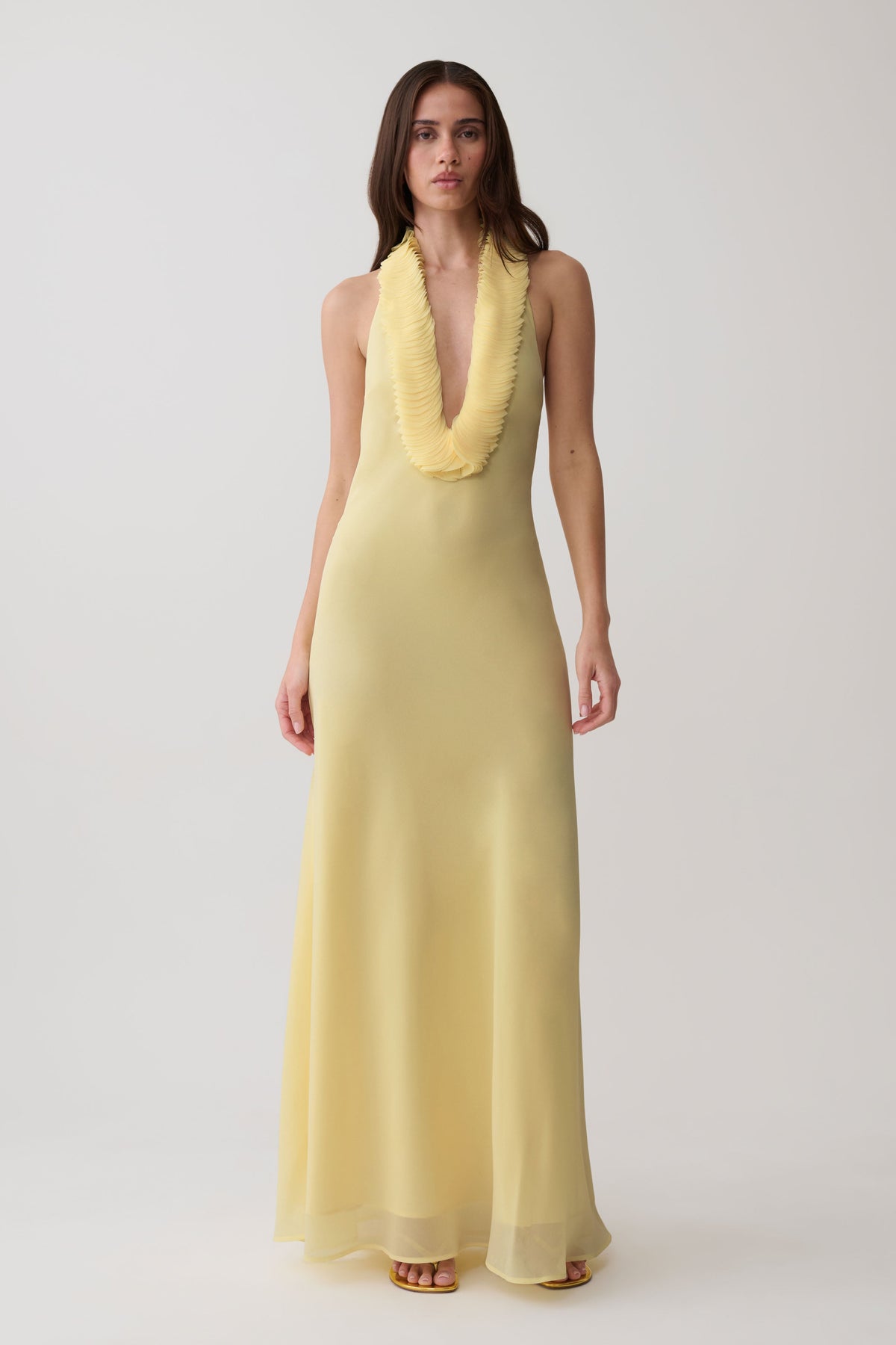 A woman modeling a sleeveless pale yellow maxi dress with a deep V-neckline and a textured, ruffled collar, set against a plain light gray studio background.