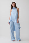 A model wearing a light blue sleeveless draped top and matching wide-leg pants, photographed against a plain light gray studio background.