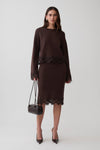 A woman models a dark brown long-sleeve knit jumper and midi skirt with dark brown lace trim, holding a small black handbag against a light gray studio background.