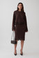 A woman models a dark brown long-sleeve knit jumper and midi skirt with dark brown lace trim, holding a small black handbag against a light gray studio background.